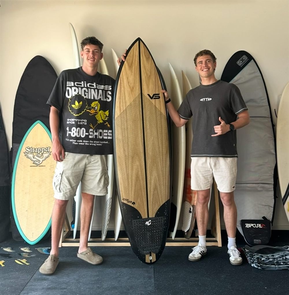 Two customers standing either side of a timber-deck surfboard they have just bought, in front of the board rack at Line Up Supply